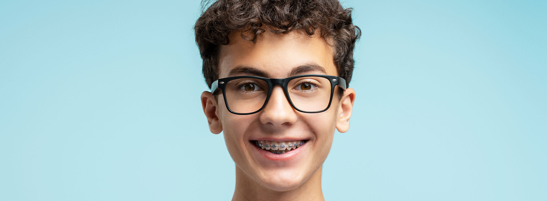 The image features a young person with glasses, smiling at the camera. They have short hair and appear to be wearing braces. The background is plain and light-colored, providing a clear contrast to the subject.