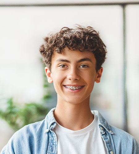 The image shows a young person with curly hair, smiling at the camera. They are wearing a blue denim jacket and have short hair. The background is blurred but appears to be an indoor setting with a window and a plant.
