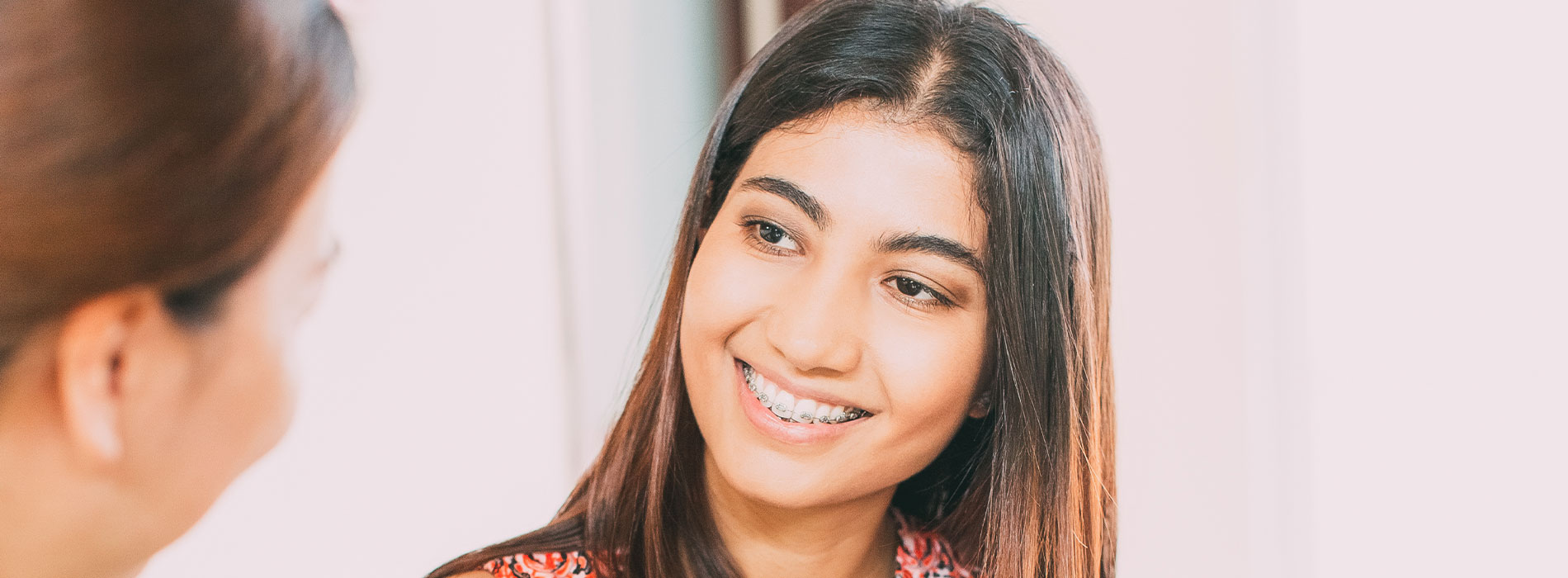 The image is a photograph of two individuals, likely engaged in a conversation or an interaction. The person in the foreground appears to be smiling and looking towards the camera, while the other individual s face is partially obscured by the first. They are both indoors with natural light illuminating their faces.