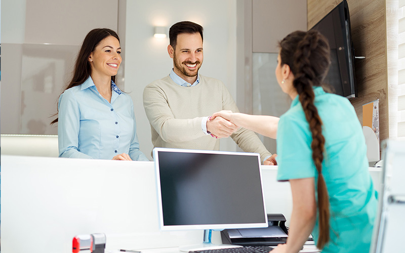 The image depicts a man and woman standing behind a reception desk, shaking hands with a person on the other side of the desk. They are smiling and appear to be engaged in a friendly interaction.