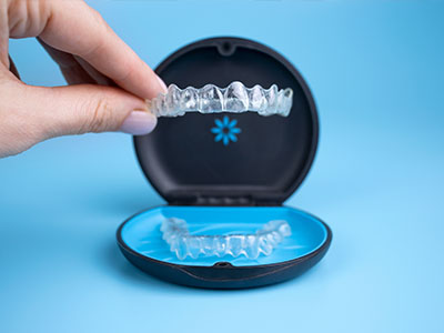 The image shows a person s hand holding an open box containing a set of transparent dental aligners, with the aligners partially inserted into a blue tray.