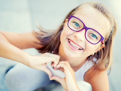 A young girl with glasses and a heart-shaped gesture, smiling in front of a blurred background.