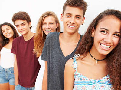 The image shows a group of four individuals, likely teenagers or young adults, posing together with smiles. They are standing in front of a plain background and appear to be at an outdoor location based on the lighting.